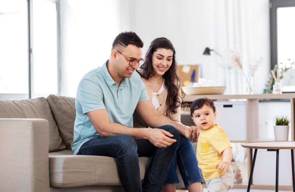 Parents on a couch smiling as their toddler stands beside them